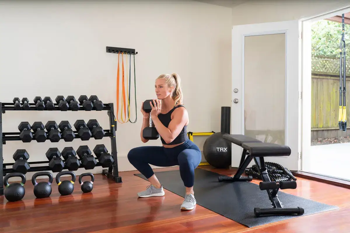 Femme faisant un squat avec haltère pour muscler son fessier. Haltères, kettlebells et bandes de résistance à proximité.