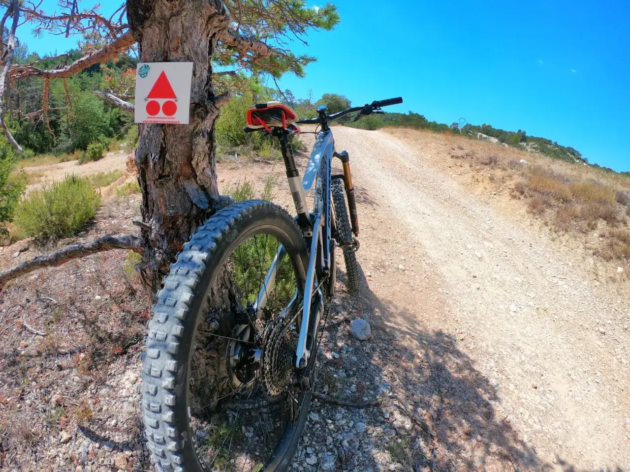 Un VTT pr&ecirc;t pour une balade sur un sentier rocailleux sous un ciel bleu. Un panneau de balisage indique la piste.