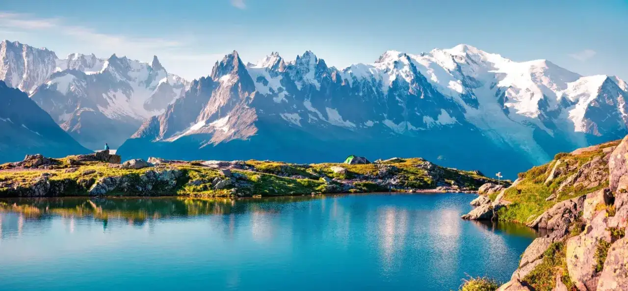 Lac alpin paisible reflétant des montagnes enneigées. Un campement discret suggère la difficulté du Tour du Mont Blanc en 7 jours.