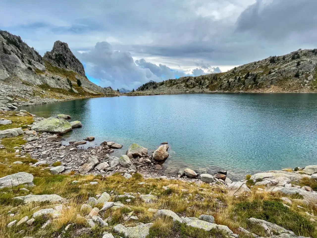 Lac d'altitude aux eaux bleues, entouré de rochers et d'herbes sèches. Un paysage typique du tour du Mercantour sous un ciel nuageux.