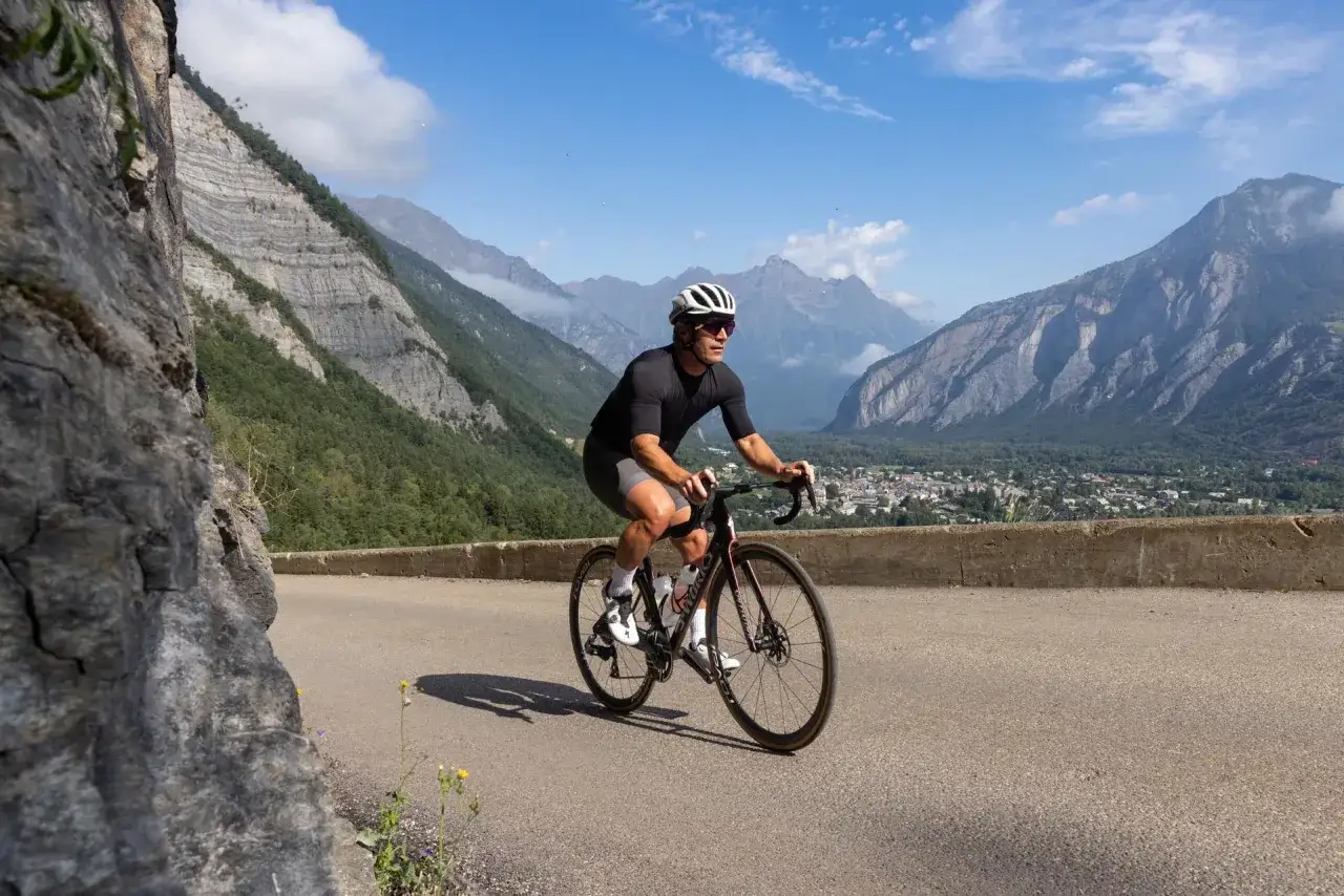 Cycliste en plein entrainement v&eacute;lo, gravissant une route de montagne spectaculaire avec un village en contrebas et des sommets majestueux.
