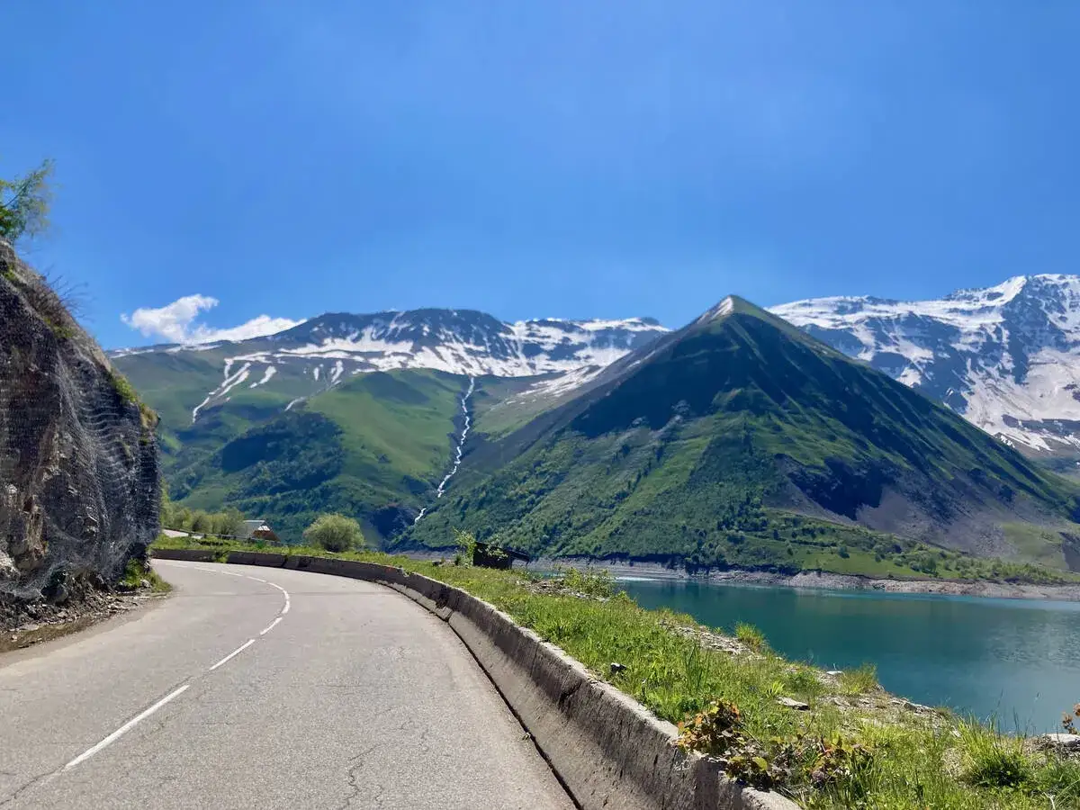 Route sinueuse menant au col du Glandon, avec un lac turquoise et des montagnes enneigées en arrière-plan.