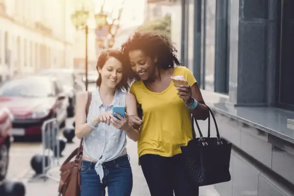 Deux amies marchent en ville, l'une montre son téléphone, l'autre mange une glace. Elles atteignent sûrement leurs 10000 pas par jour !