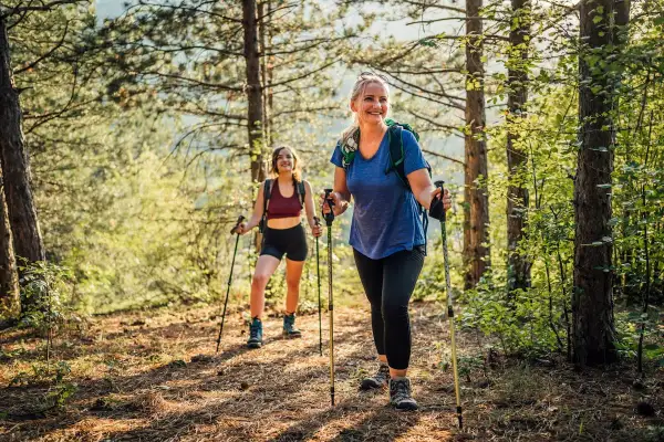 Deux femmes souriantes randonnent en forêt avec des bâtons, profitant des marche avec bâton bienfaits pour la santé et le bien-être.