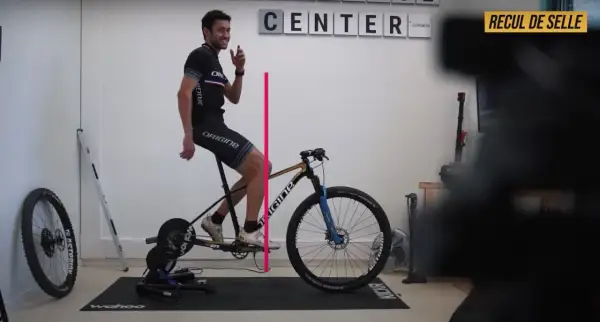 Un cycliste ajuste le recul de selle de son vélo sur un home trainer. Une ligne rose indique la mesure.