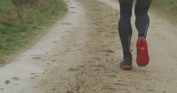Jambes d'un coureur sur un chemin de terre, concentré sur sa cadence course à pied. Les chaussures rouges et noires s'enfoncent dans le sol humide.