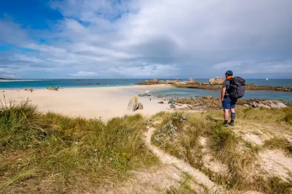 Randonneur contemplant une plage de sable fin, des rochers et la mer. Le sentier GR34 serpente dans les dunes.
