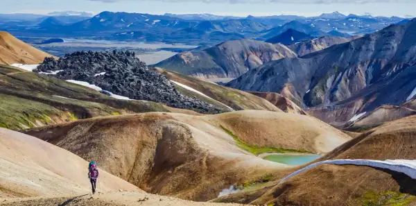 Randonneuse solitaire explorant les paysages colorés et volcaniques du trek Landmannalaugar, avec des montagnes aux teintes ocre, vertes et bleues.