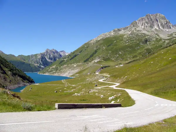 Route sinueuse menant au col du Glandon, avec un lac bleu vif et des montagnes verdoyantes sous un ciel clair.
